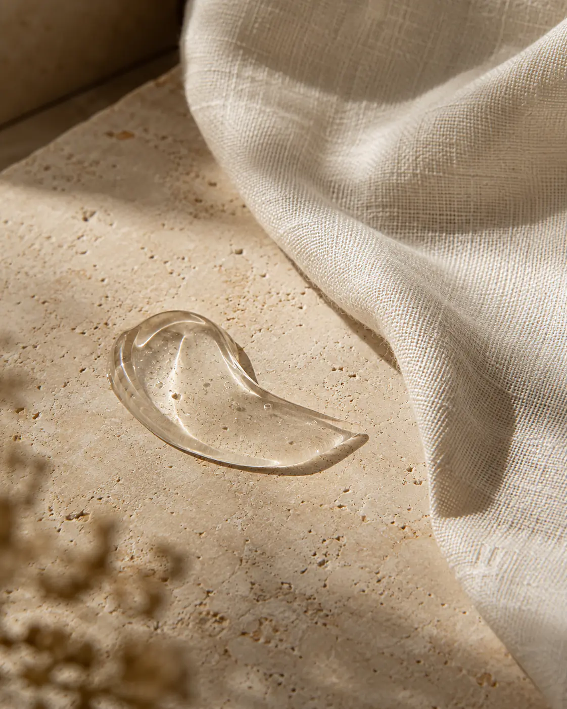 Translucent skincare gel swatch beside ivory linen on travertine.